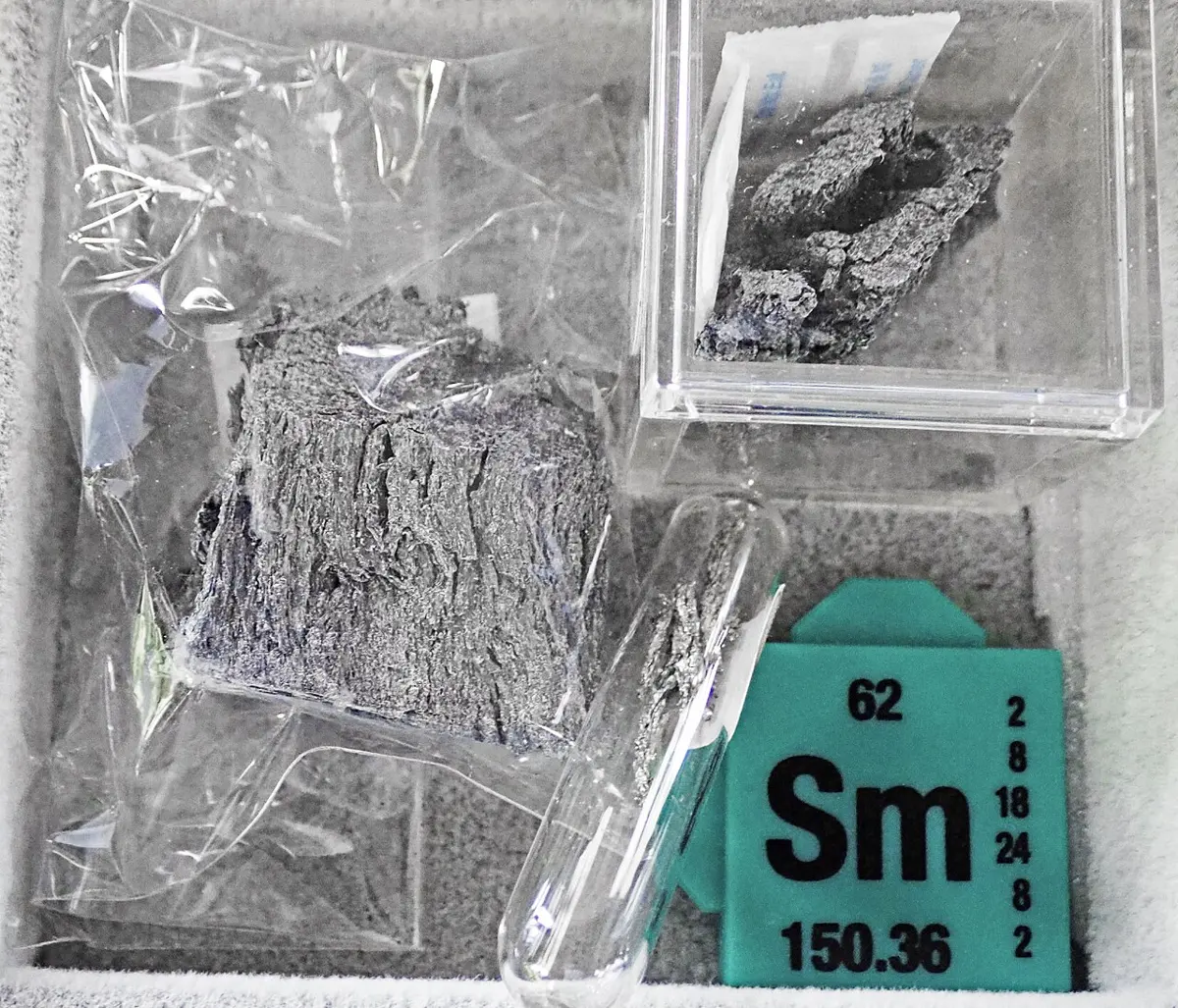 Gray, flaky pieces of metal in plastic bags and a small clear box, beside a green card that reads Sm, 62