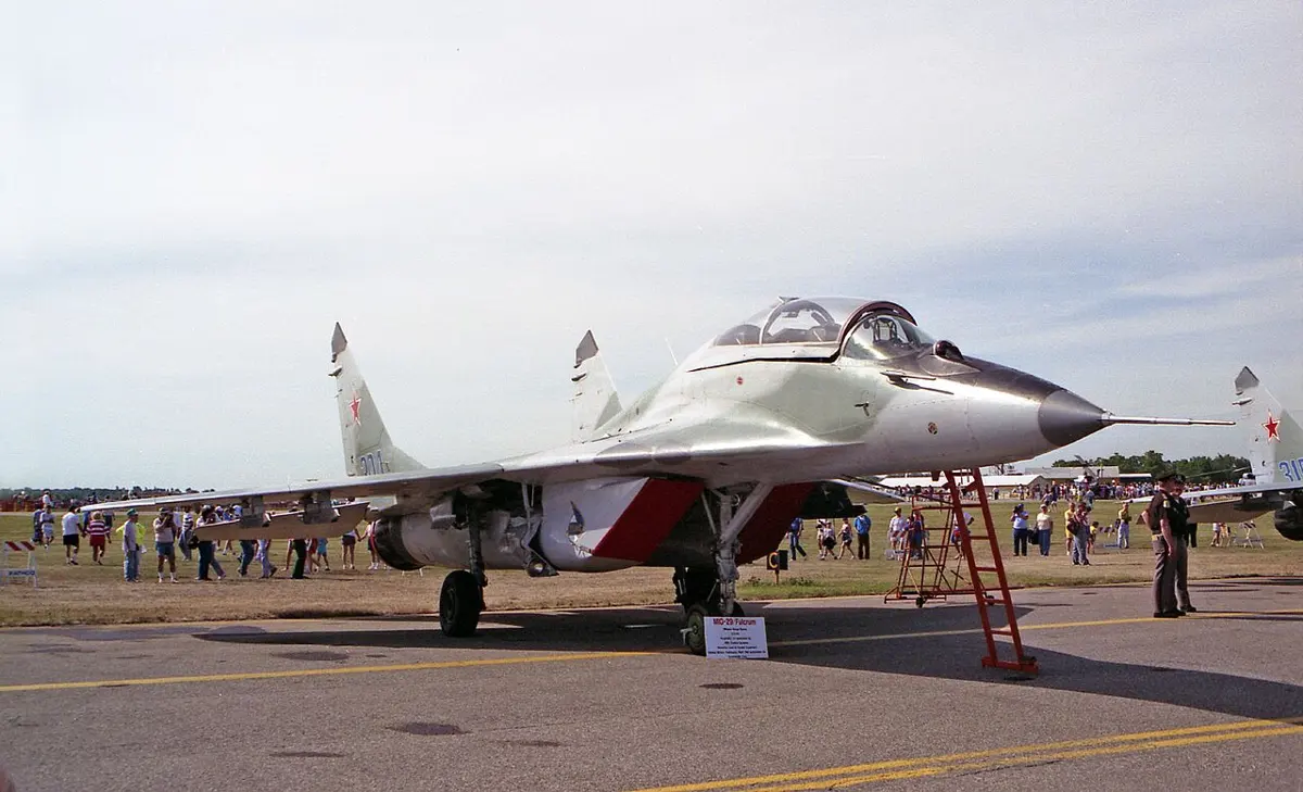 A gray jet fighter with two tail fins and a red star, parked on a runway at an air show, with people walking around it