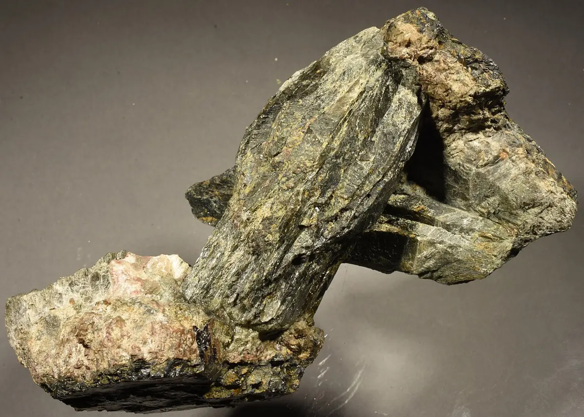 A rock specimen with long, dark gray-green crystals rising from a pale pinkish base