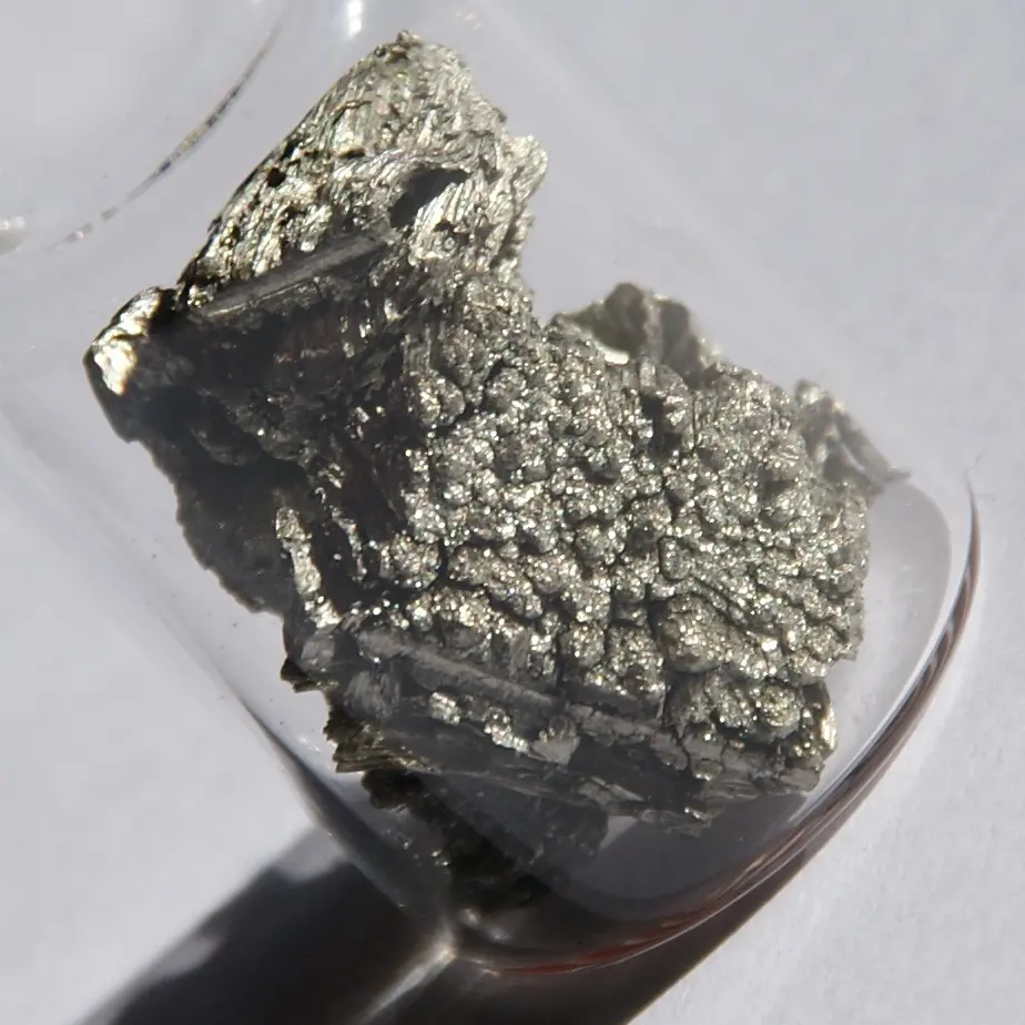 A rough, sparkling silver-gray lump of metal crystals in a small glass vial