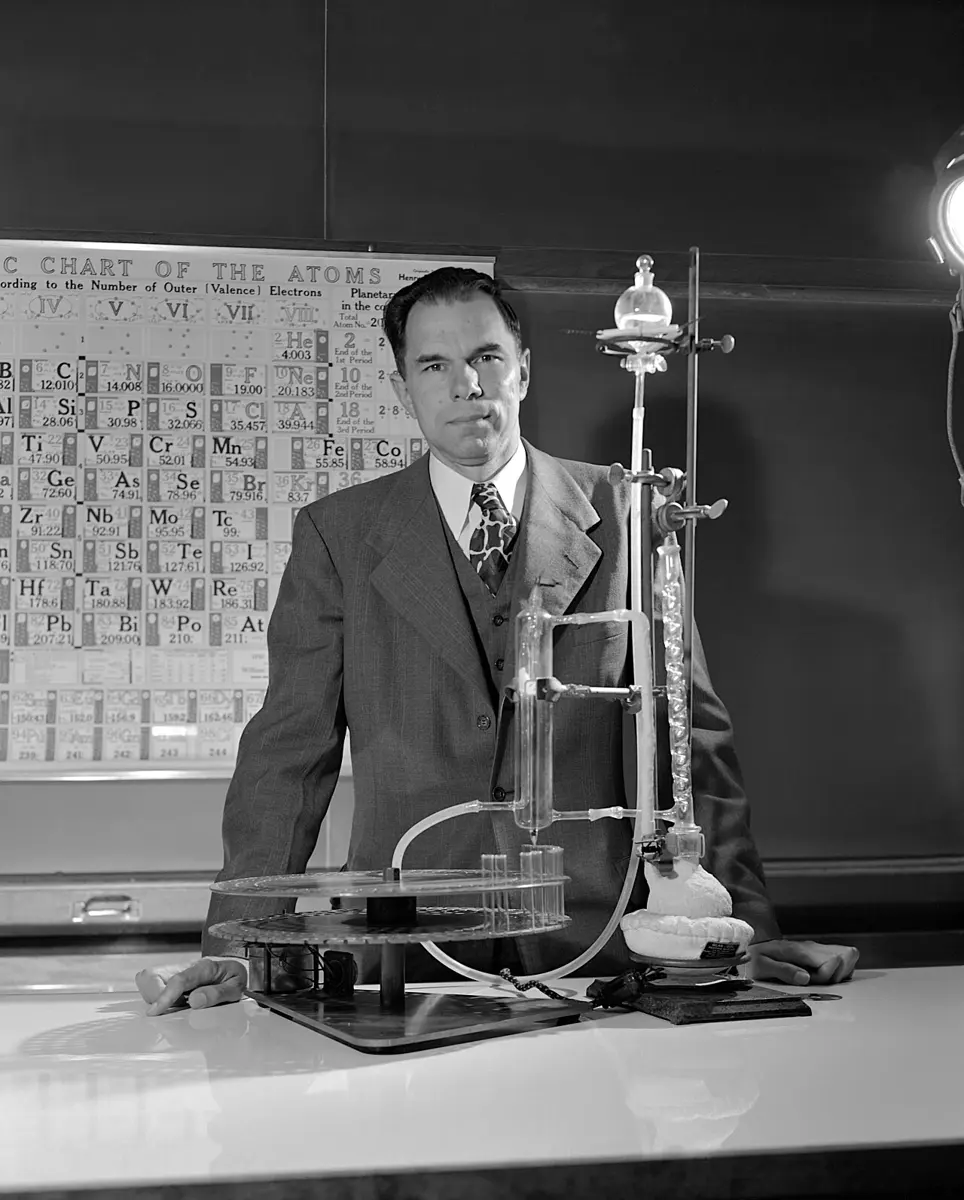 A black-and-white photo of a man in a three-piece suit standing at a laboratory bench beside a tall glass column held in a metal stand, with a large chart of the elements on the wall behind him