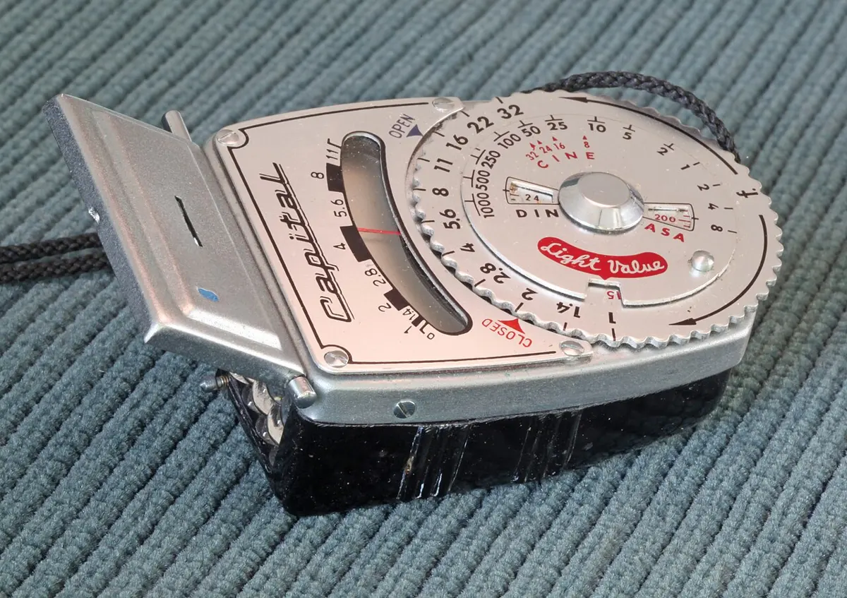 An old silver handheld light meter with a round dial marked with numbers and a window at the front, lying on a blue cloth