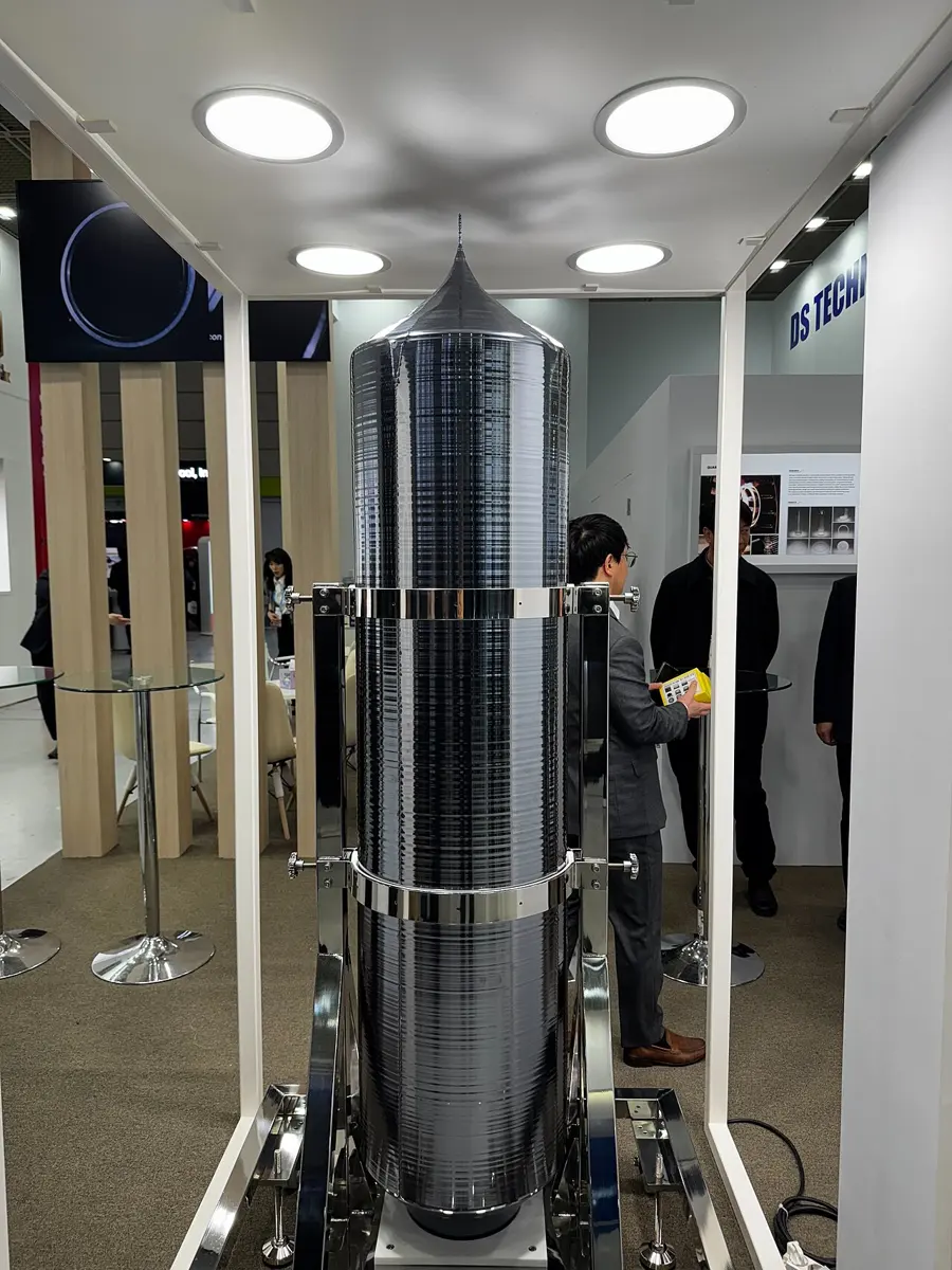 A tall, dark gray, shiny cylinder with a pointed top, taller than the man standing beside it, held upright in a steel frame at an exhibition