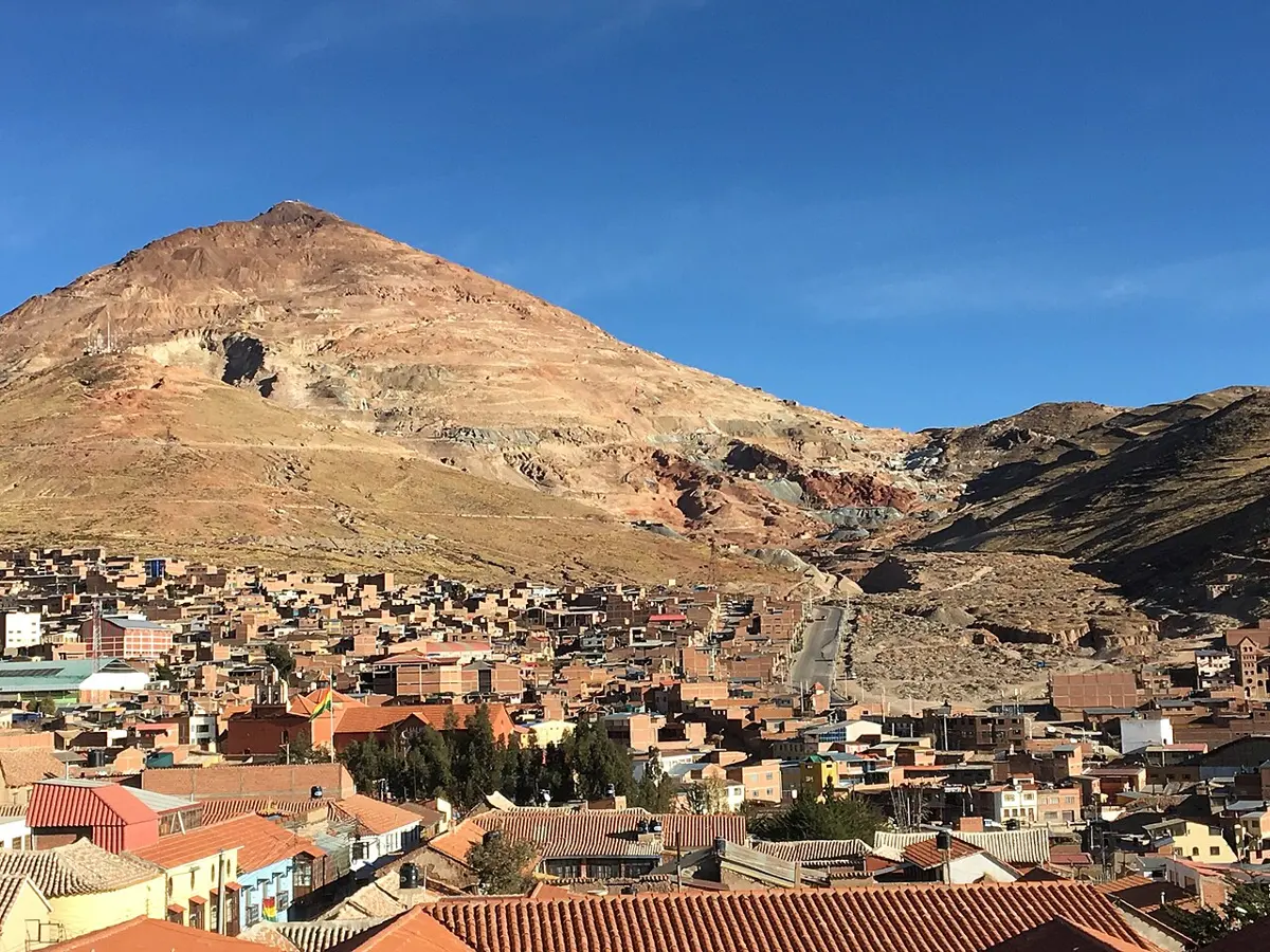 A large reddish-brown cone-shaped mountain rising behind a town of red-roofed houses under a blue sky