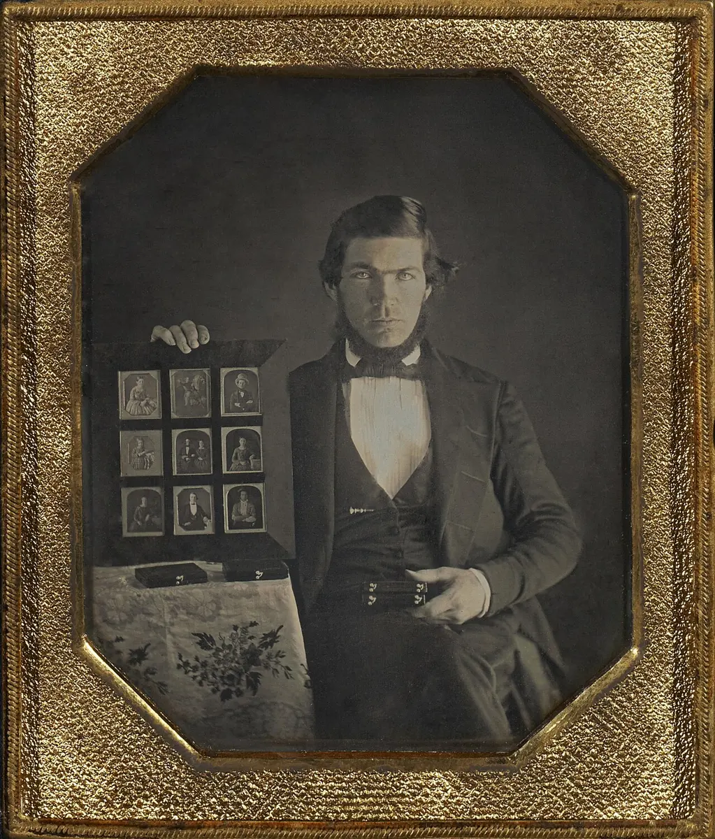 An old brownish photograph in a gold-colored frame of a young man in a dark coat holding up a board of nine small framed photographs