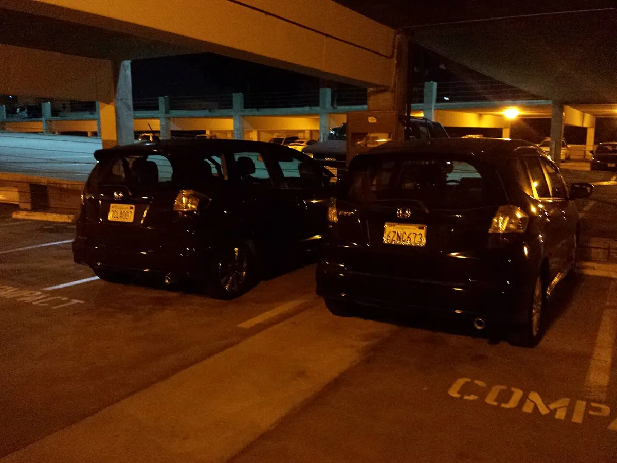 Two small hatchback cars side by side in a parking garage under yellow-orange lamps; both look black