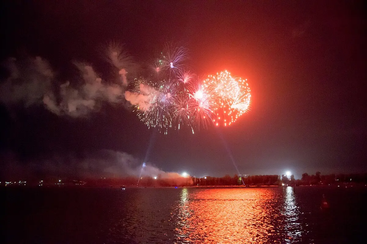 Red fireworks bursting in a night sky over a river, with white sparkling fireworks beside them and smoke drifting to the left