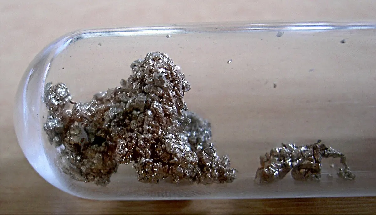 Rough, sparkling lumps of gray-gold metal crystals lying in a sealed glass tube