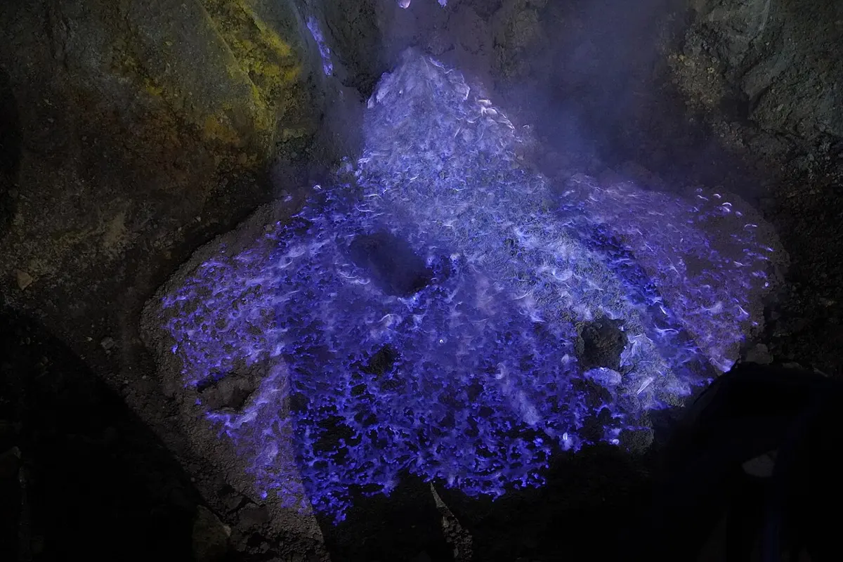 Bright violet-blue flames spreading over dark rocks at night, with pale smoke rising above them