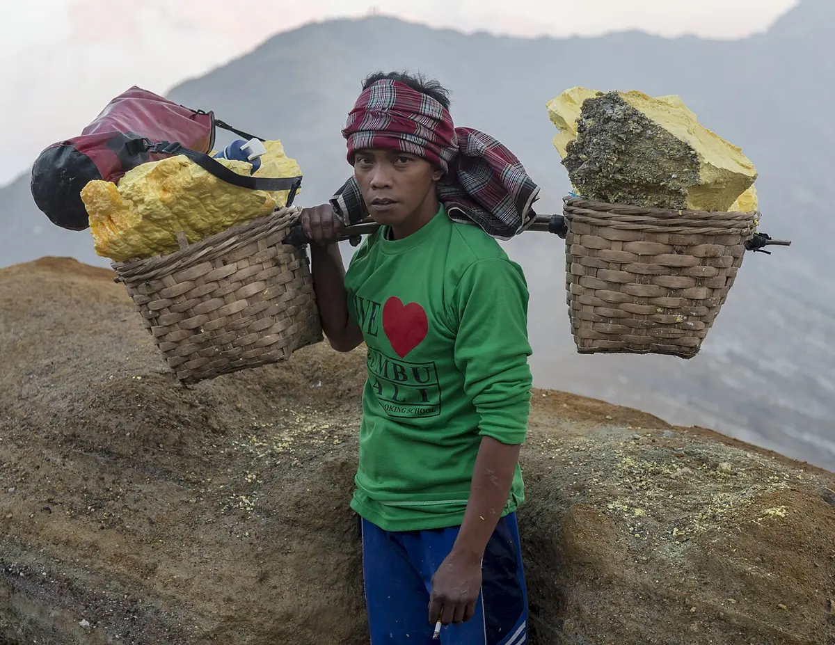 A young man in a green T-shirt and a checked headscarf, with two woven baskets of big yellow sulphur lumps hanging from a pole across his shoulder, standing on a rocky crater rim