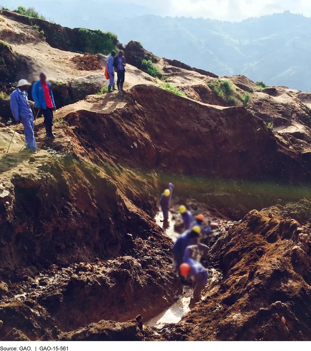 Miners in blue overalls and hard hats digging in a muddy, water-filled trench cut into a reddish-brown hillside, while others stand watching at the top