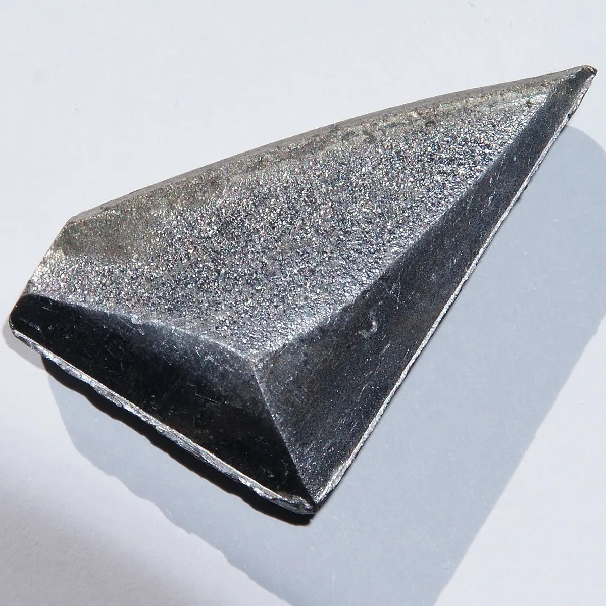 A wedge-shaped lump of dark gray metal with a rough, sparkling top and smooth, shiny sides, on a white surface