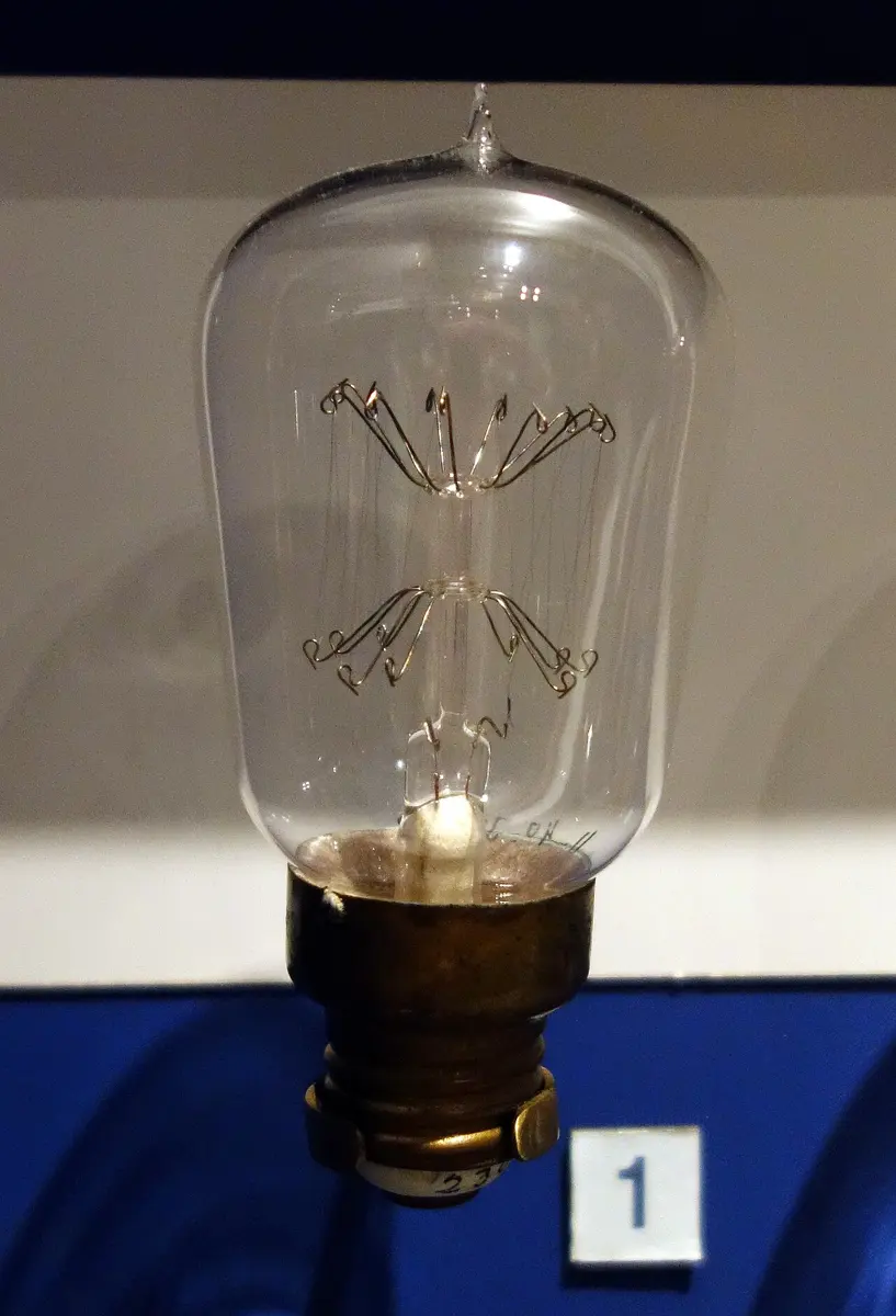 An old clear glass bulb on a brass screw cap; inside, a long, thin wire zigzags up and down between two star-shaped rings of hooked supports on a glass rod