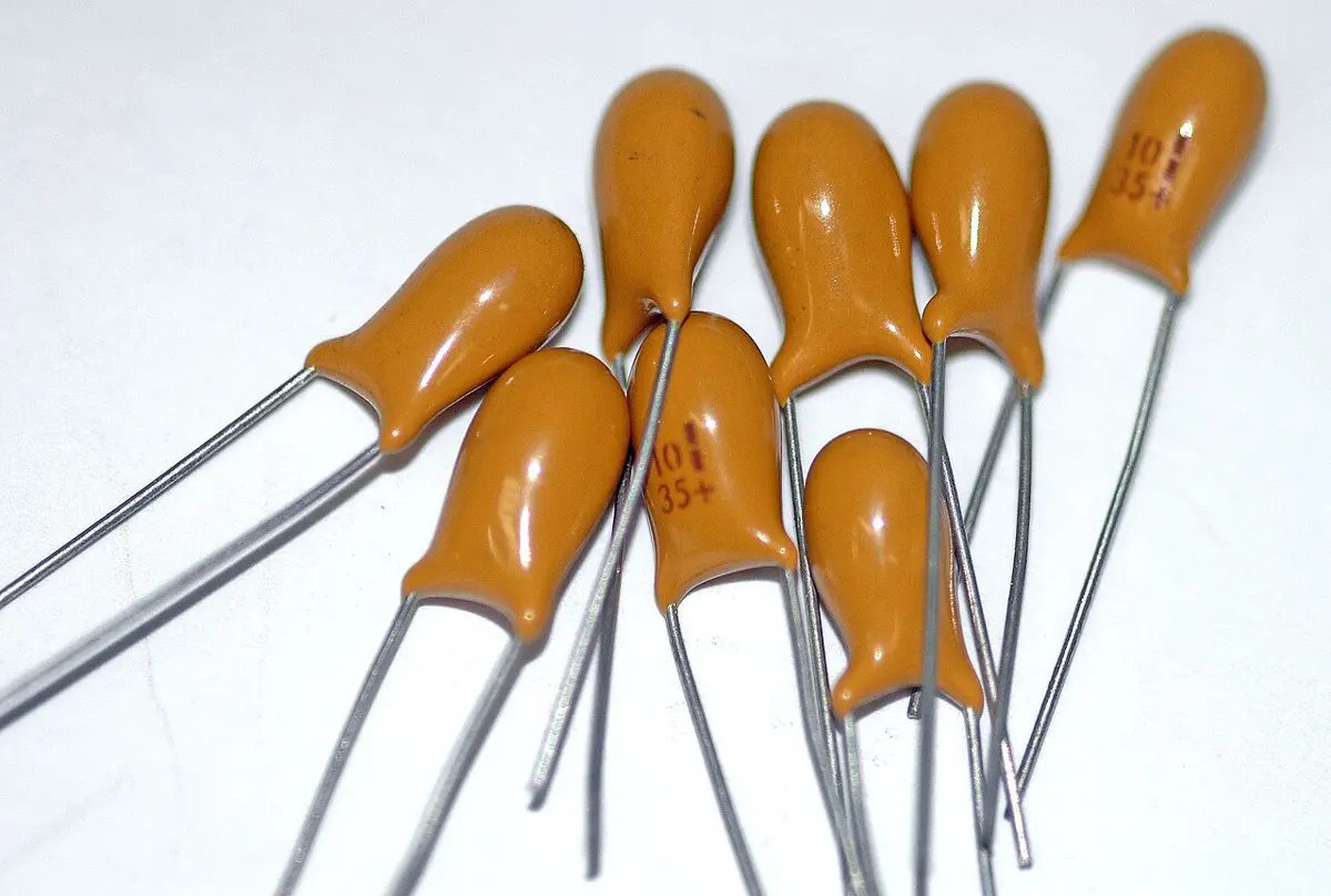 Eight small orange, teardrop-shaped electronic parts, each with two thin silver wire legs, lying on a white table; two are printed with 10 and 35+