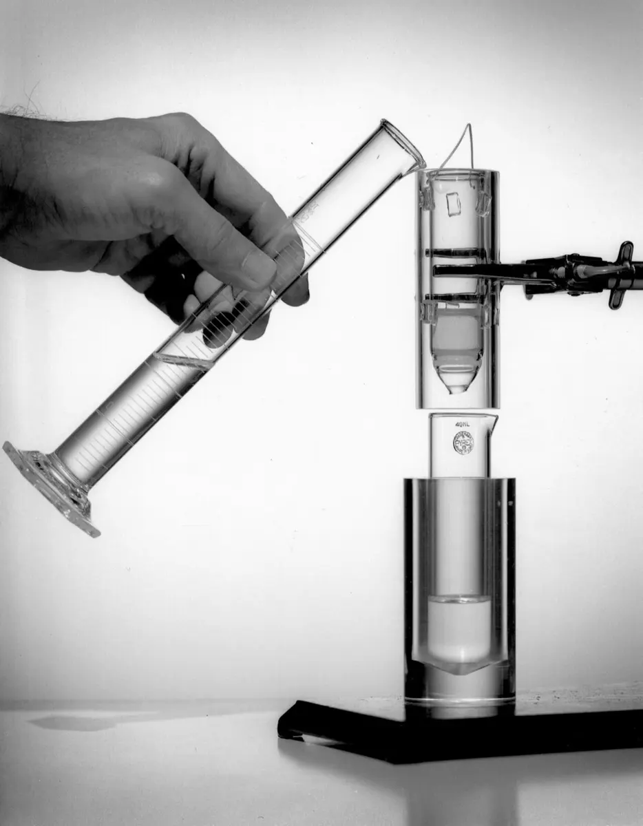 A black-and-white photograph: a hand pours liquid from a measuring cylinder into a glass column clamped upright, which drips into a small bottle standing in a clear holder below