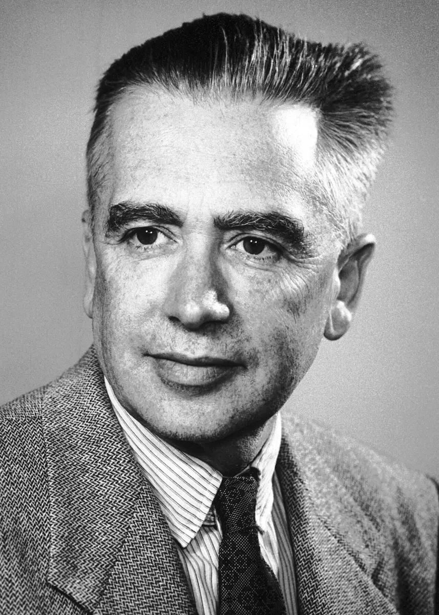 A black-and-white portrait photograph of a man with graying hair brushed up, wearing a tweed jacket, striped shirt and patterned tie, looking at the camera