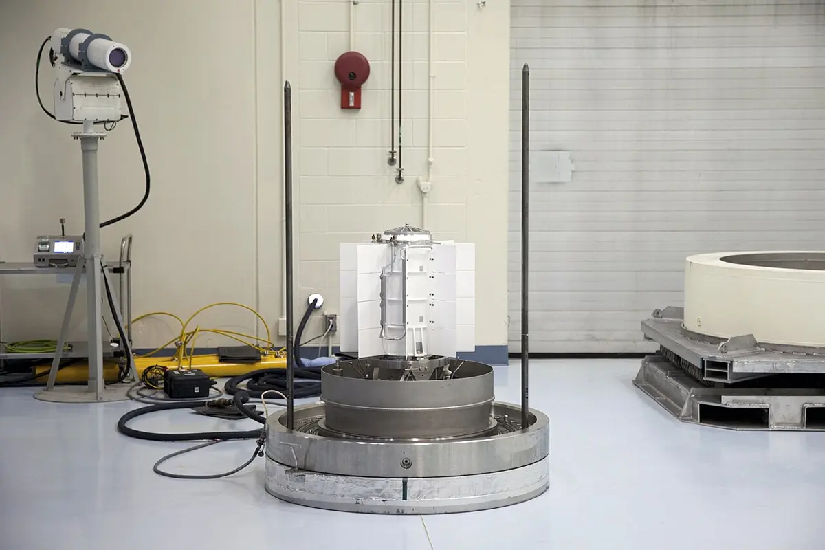 A white cylinder with flat fins around its sides, standing on a round metal base in a large, bright room, with cables on the floor