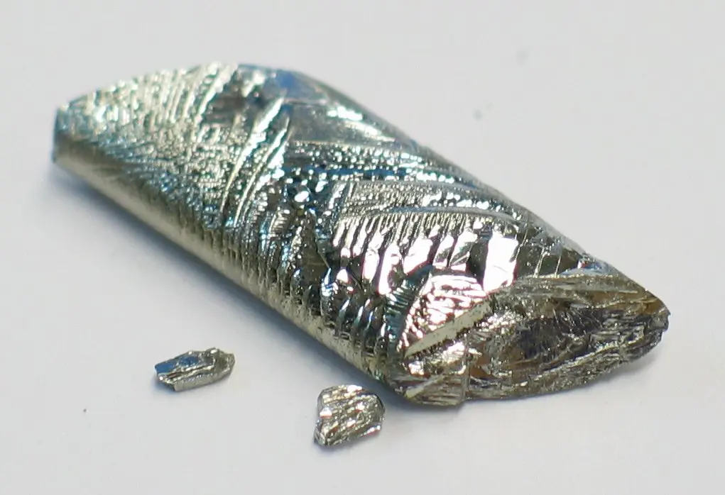 A shiny, silvery-white crystal shaped like a small bar, with a pattern of ridges on top and two tiny chips beside it