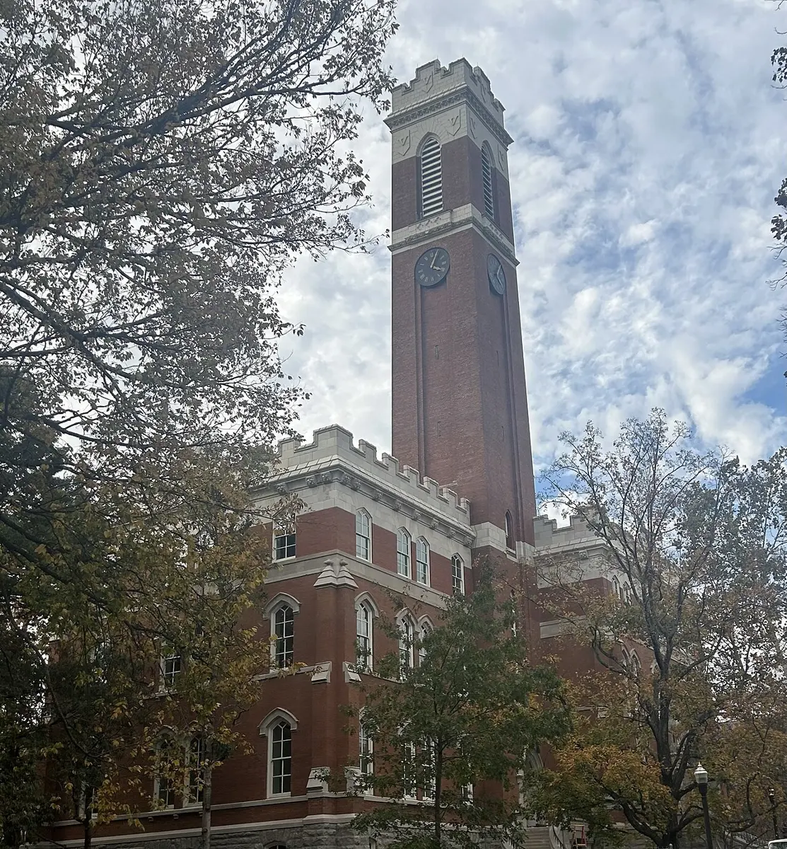 A red-brick building with a tall square clock tower, seen from below through autumn trees against a cloudy sky