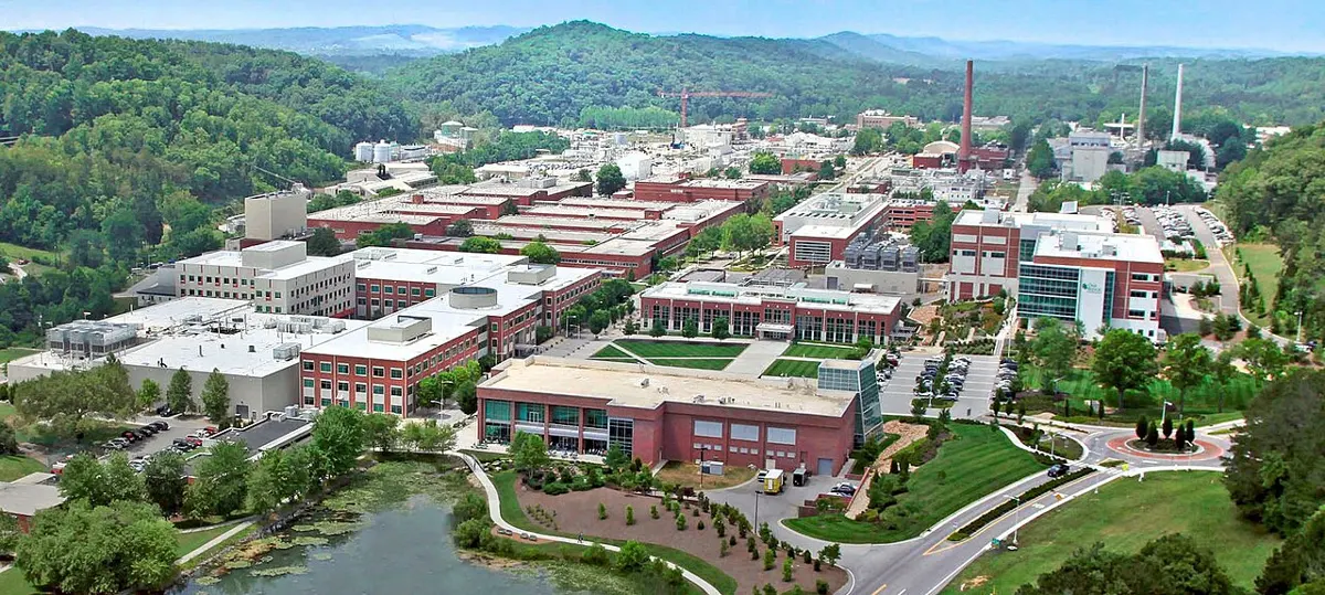 An aerial view of a large campus of modern brick buildings, lawns and a pond, surrounded by wooded hills