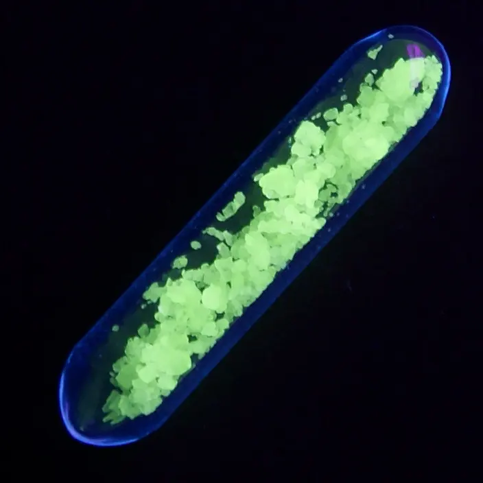 A glass tube in the dark, full of small crystals glowing bright yellow-green