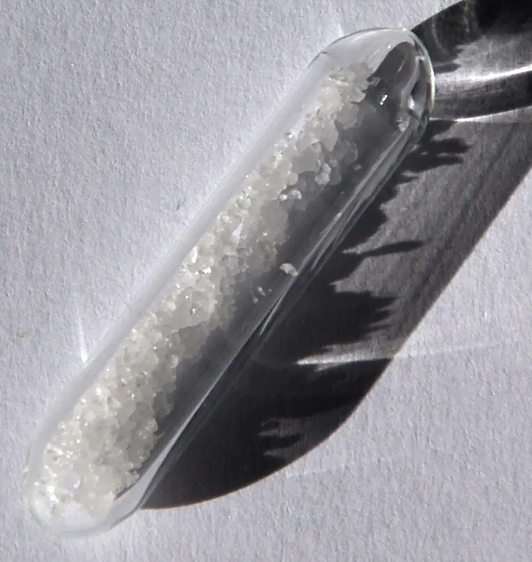 A clear glass tube, lying on a white surface in sunlight, half full of small white crystals