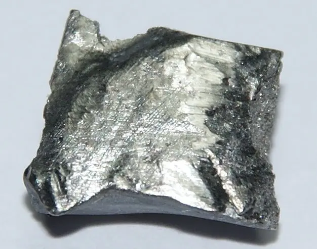 A flat, roughly square piece of shiny silver-gray metal with a ridged, partly dark surface