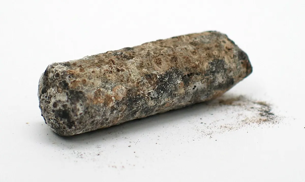A short rod lying on a white surface, covered in a rough crust of gray, brown and black, with dark powder flaking off beside it