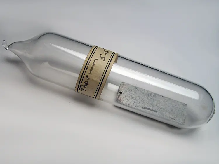 A sealed glass tube with a handwritten paper label reading Thorium, holding a small flat strip of dull, speckled gray-white metal