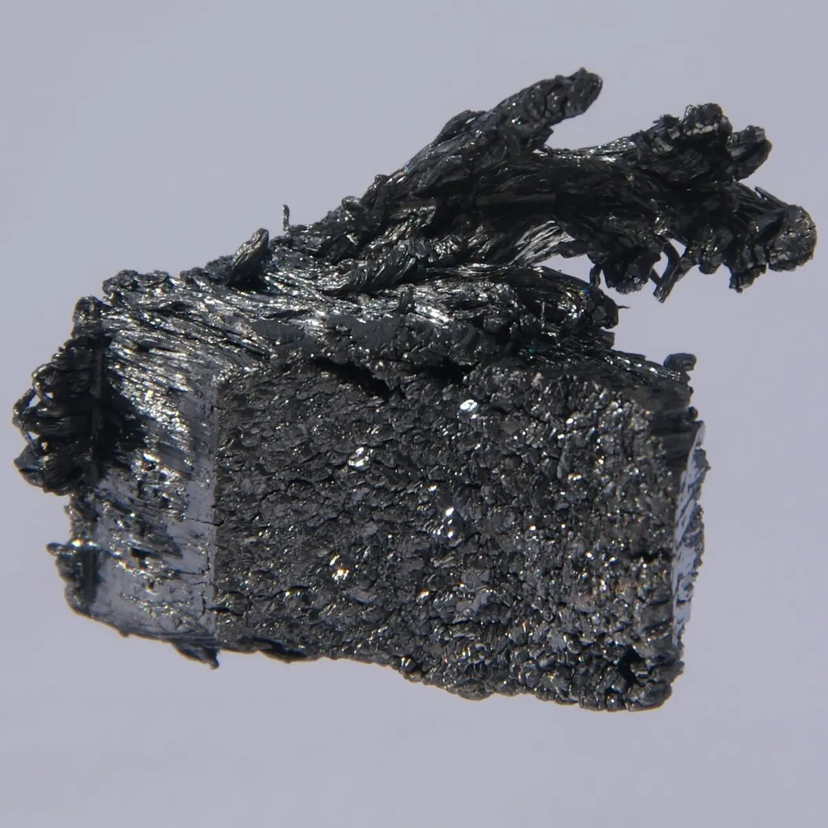 A dark gray, glittering block of metal crystals, with a feathery tuft of thin needle-like crystals growing from its top