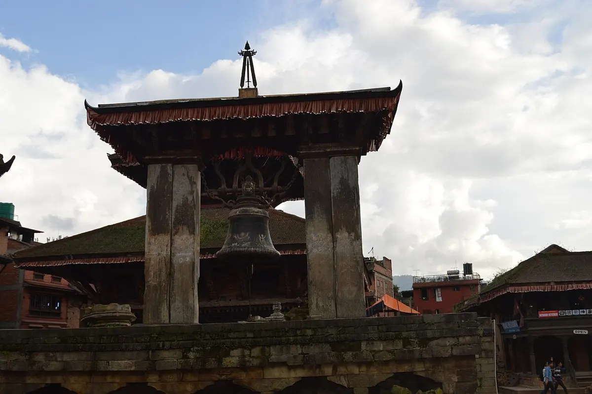 A large dark metal bell hanging under a small tiled roof held up by two thick stone pillars, with temple roofs behind and a cloudy sky