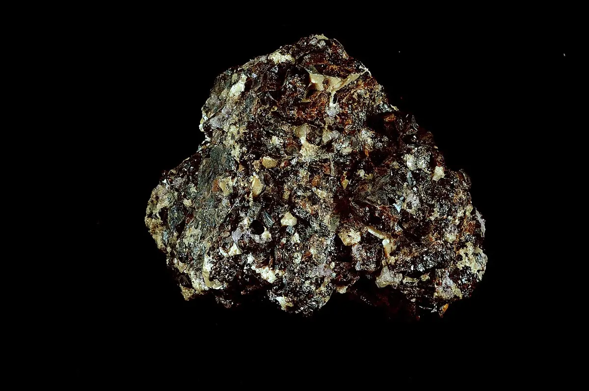 A lumpy rock on a black background, made of many small, shiny, dark brown to black crystals with a few pale yellowish ones between them