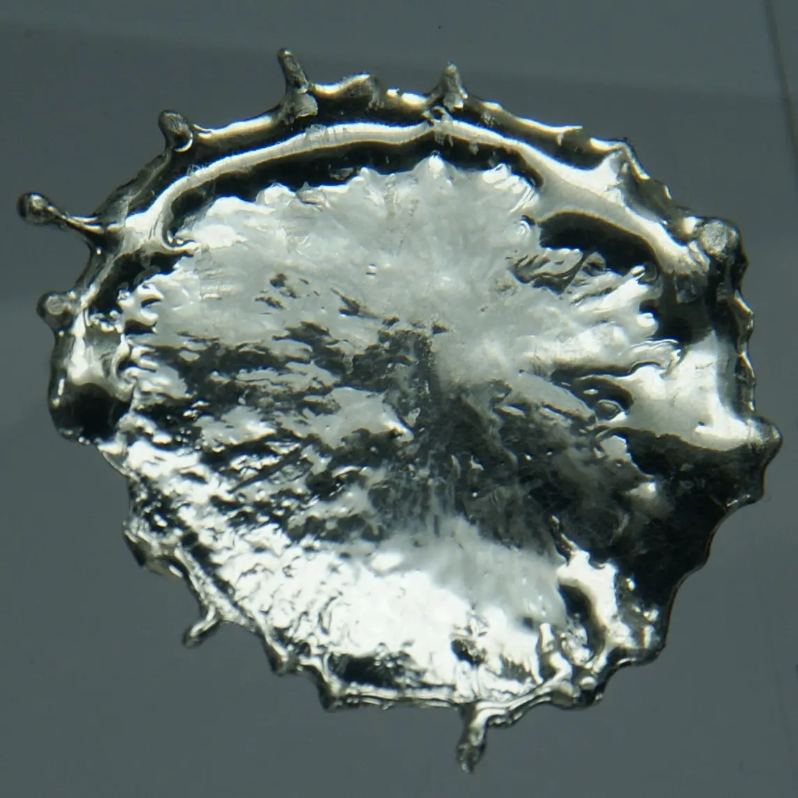 A flat, round splash of bright silvery metal with a rippled surface and little spikes round its edge, on a gray background