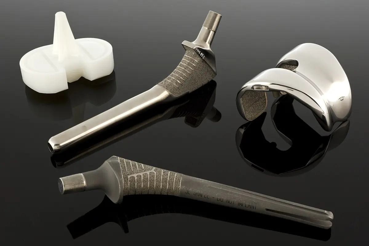 Parts of an artificial hip and knee on a black surface: two long metal stems with ridged upper parts, a curved shiny metal piece and a white plastic piece