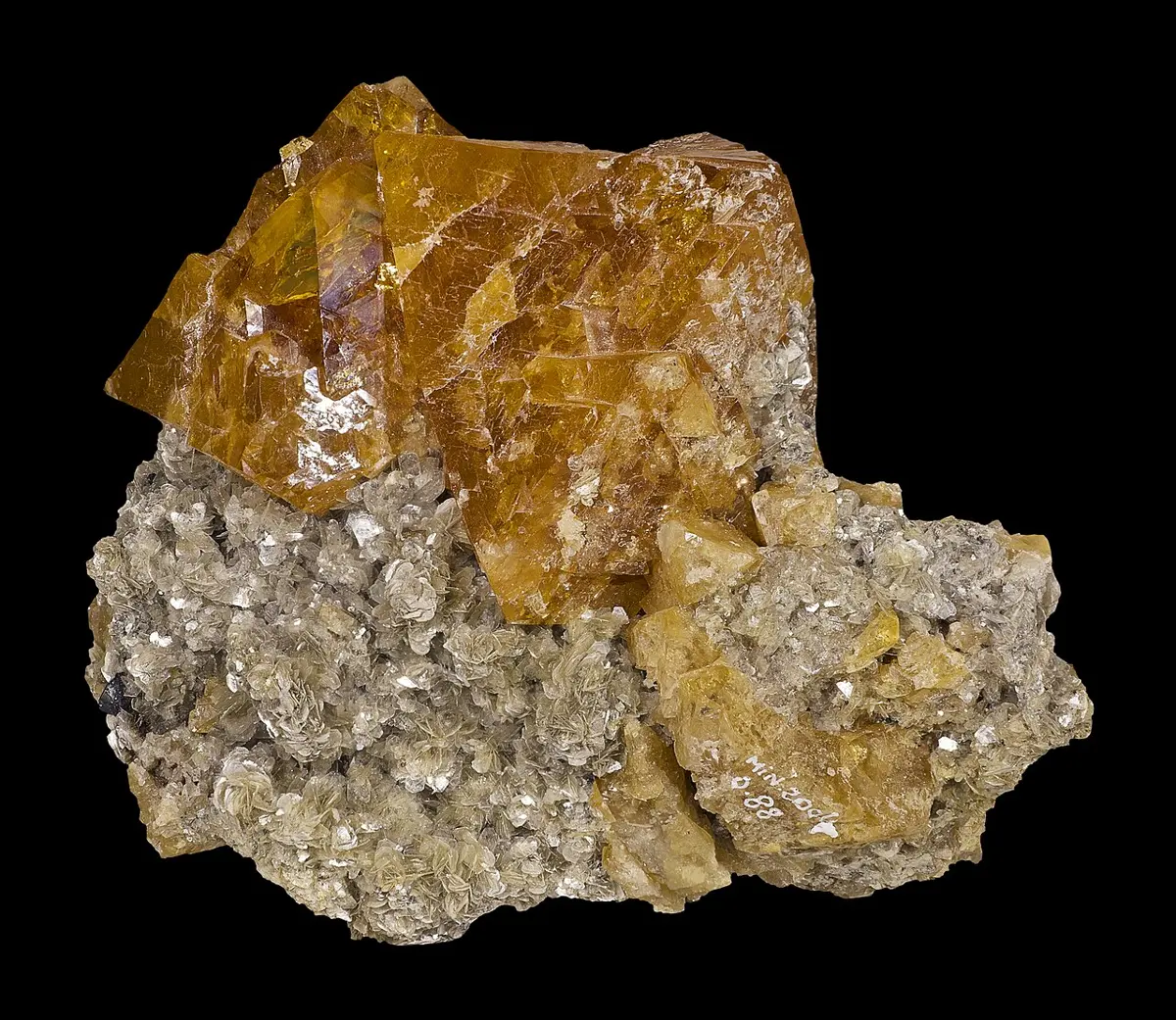A rock with a large, glassy, honey-orange crystal sitting on a bed of small, shiny, silvery-gray flakes