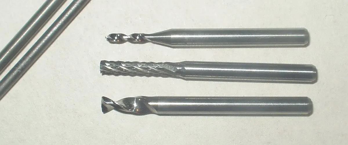 Three small drill bits and cutters with silvery shafts lying side by side, with two thin gray rods at the left