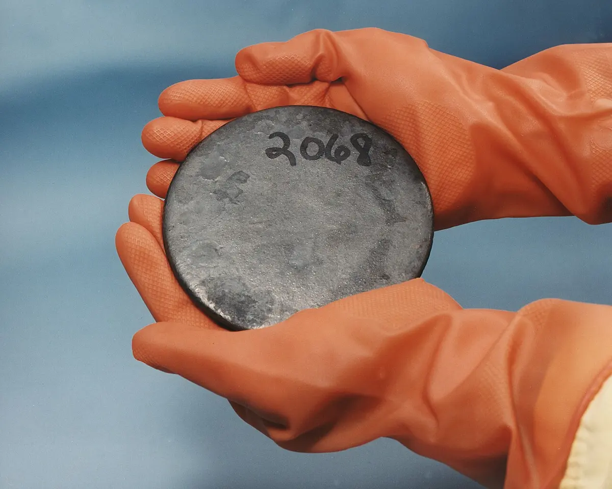 A flat, dull gray metal disc with a number written on it, held in two hands in orange rubber gloves