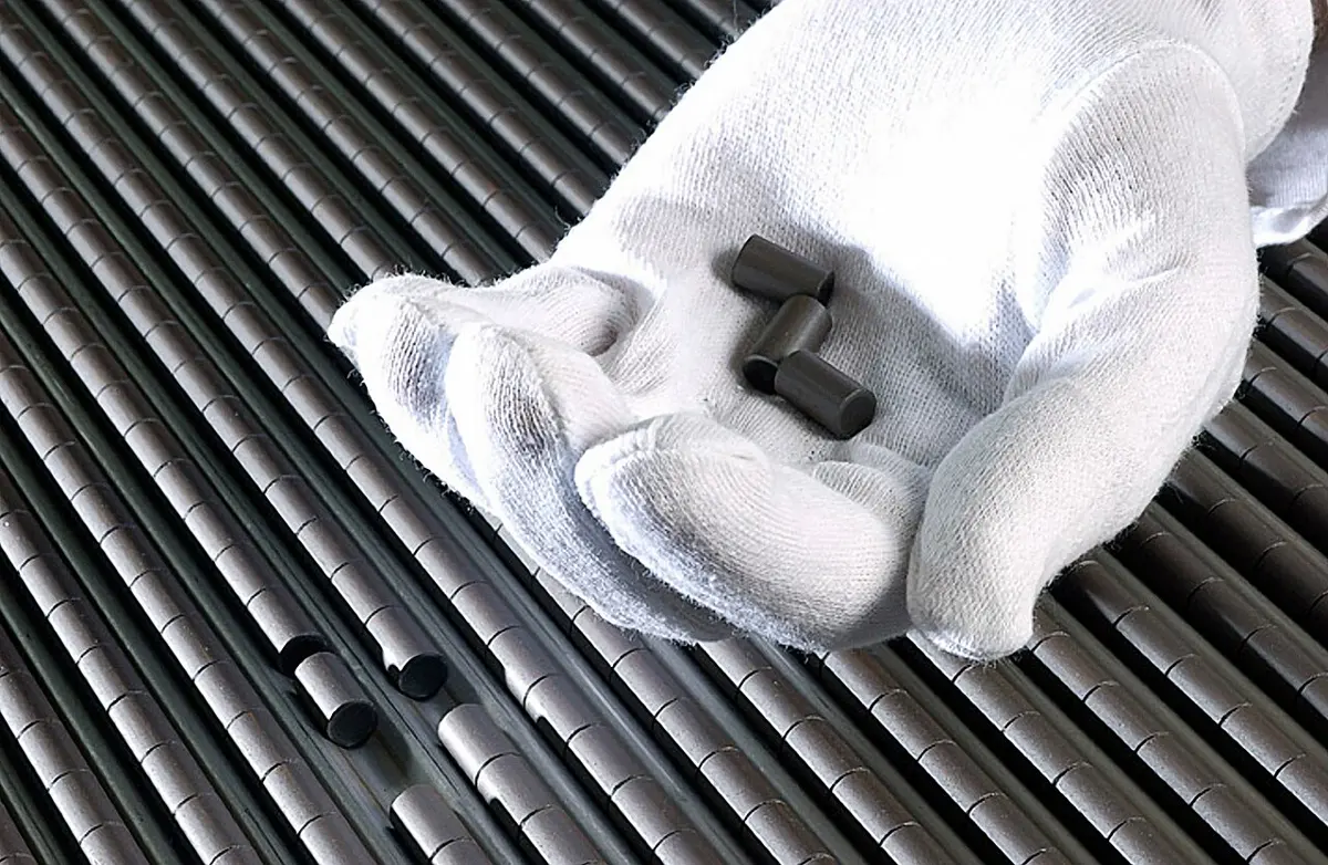 A hand in a white glove holding three small dark gray cylinders, above a row of long metal tubes