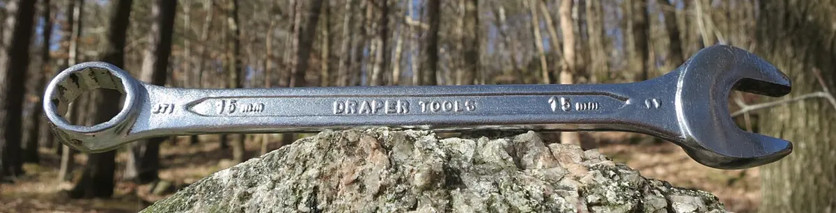 A shiny steel spanner resting on a rock in a wood, ring-shaped at one end and open at the other, stamped 15 mm and Draper Tools