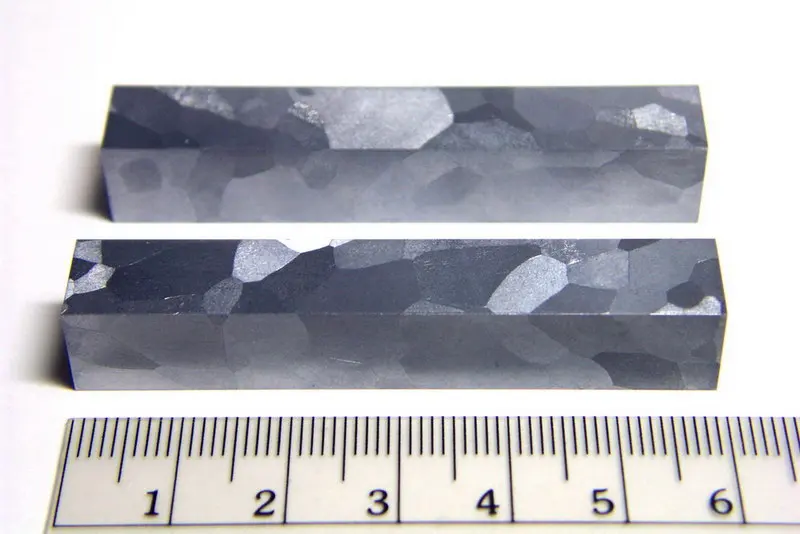 Two square bars of gray metal lying beside a ruler, their surfaces a patchwork of lighter and darker patches