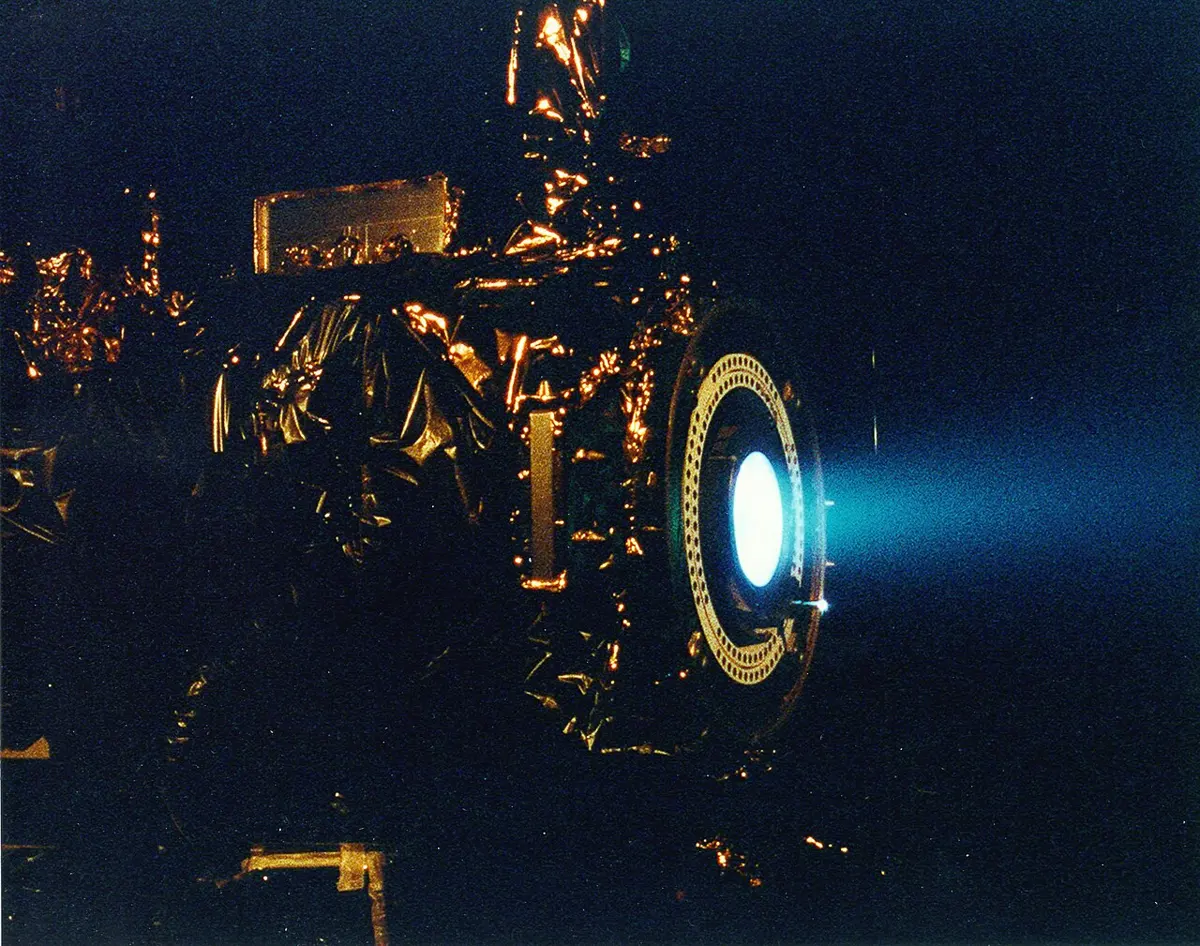 An engine wrapped in gold foil in a dark chamber, with a round opening glowing bright blue and a pale blue plume streaming out of it