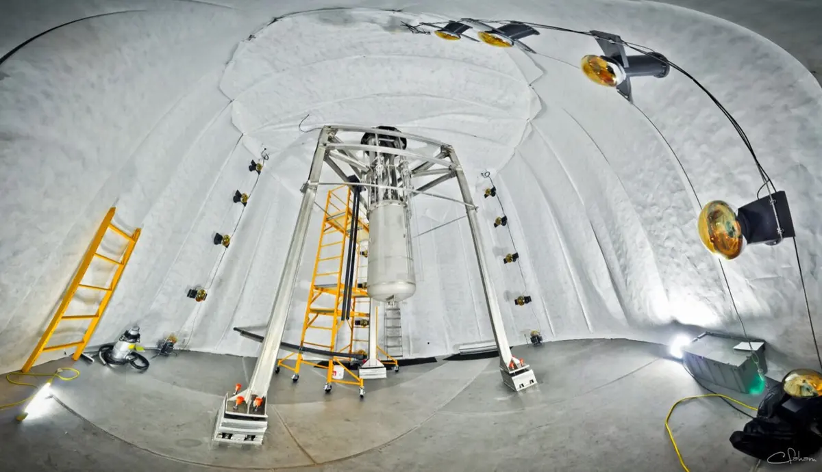 Inside a huge white-lined tank, a tall shiny metal cylinder hanging in a steel frame, with ladders and lamps around it