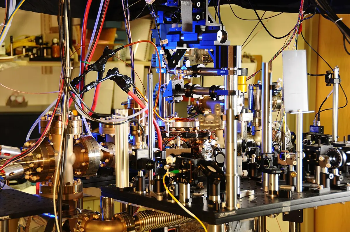 A crowded laboratory bench: metal tubes and round flanges with bolts, mirrors on posts, blue mounts, and many colored cables and optical fibers