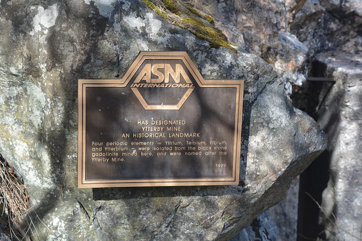 A bronze plaque on a mossy rock, headed ASM International, saying the Ytterby mine is a historical landmark where four elements, yttrium, terbium, erbium and ytterbium, were isolated from the black stone gadolinite