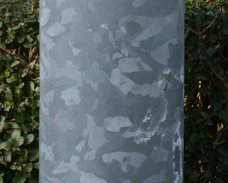 A gray metal pole in front of a hedge, its surface covered with irregular patches of lighter and darker gray