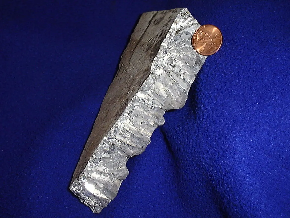 A long, rough, silvery-gray bar of metal with a crystalline surface on blue cloth, with a small copper-colored coin at its top end