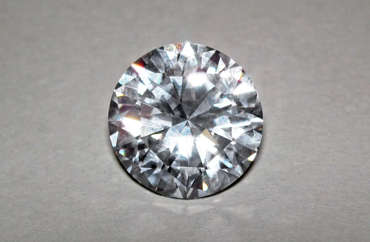 A round, sparkling, colorless cut gemstone with many facets, lying on a gray surface