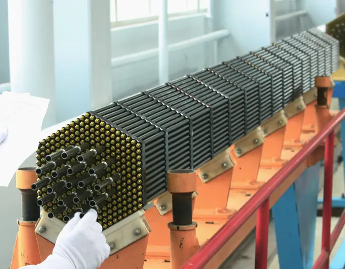 A long bundle of many thin metal rods held together in a six-sided frame, lying on an orange stand in a factory; a gloved hand touches its end, where the round ends of the rods show