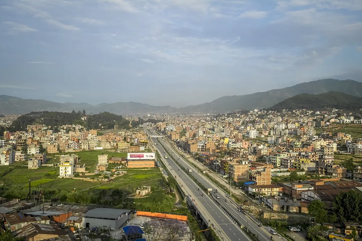 A wide, straight divided highway seen from above, running through rows of brick houses and green fields towards hills
