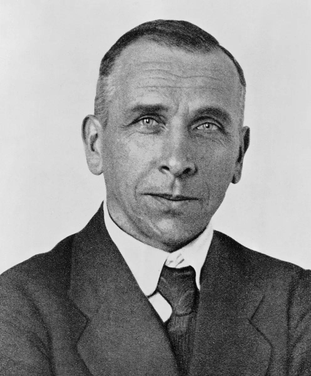 A black-and-white photograph of a man with short hair and a mustache, in a jacket and tie, looking to one side